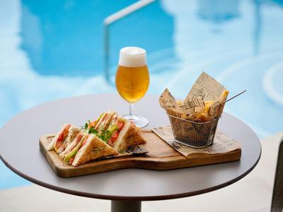 Sándwich y papas en una tabla de madera con un vaso de cerveza junto a la piscina.