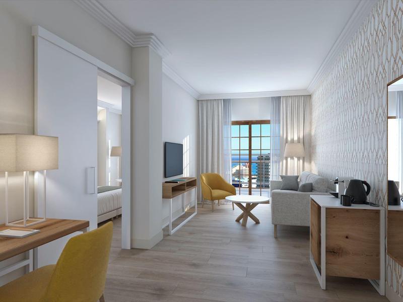 Lichte, moderne hotelsuite met zitgedeelte, gele stoelen en balkonzicht.