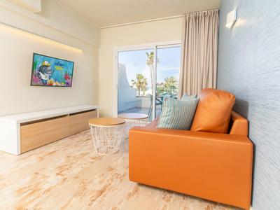 Modern hotel room with orange sofa, wall-mounted TV, wooden floor, and sliding glass door to balcony.