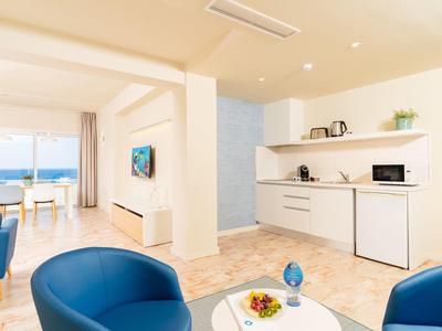 Bright, modern living space with blue chairs, white kitchenette, and dining area by large windows.