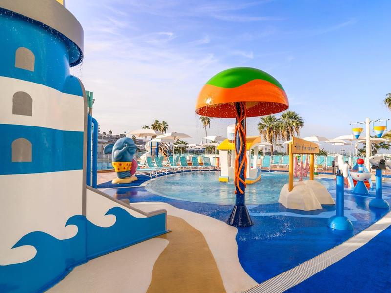 Bunter Wasserspielplatz mit planschendem Pool, Rutschen und Palmen unter blauem Himmel.
