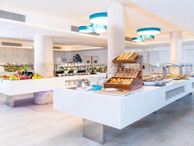 Buffet moderno de hotel con productos frescos, frutas y platos fríos en mesas blancas.