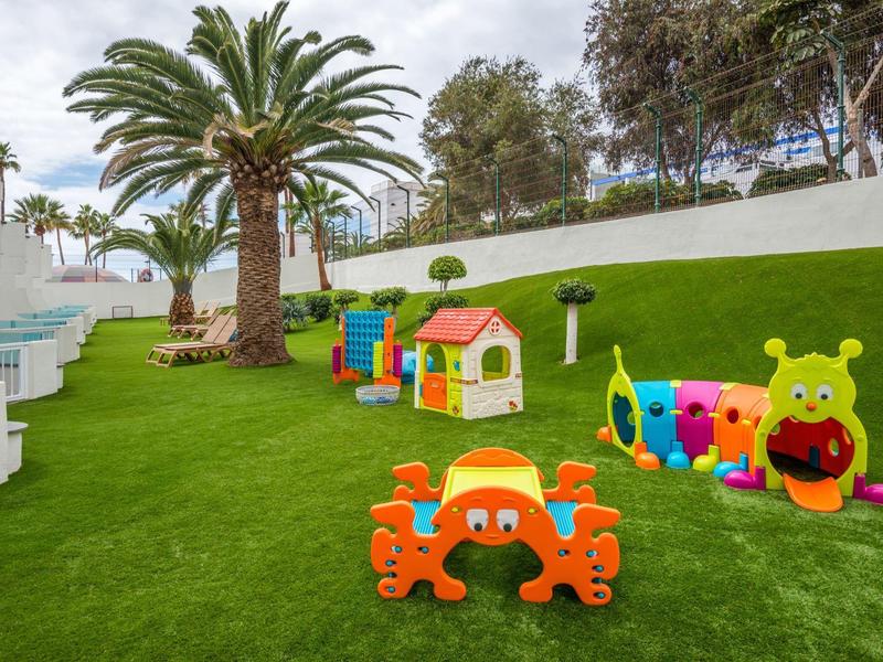 Area giochi colorata per bambini con giocattoli, prato, palma e sedute bianche vicino alla piscina.