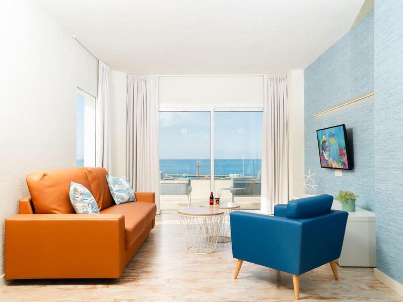 Soggiorno luminoso con divano arancione, poltrona blu, vista mare e TV su muro blu.