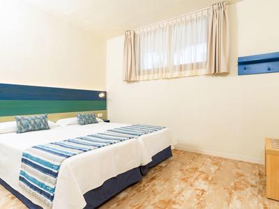 Bright hotel room with twin beds, striped bedding, window with curtains, and tiled floor.
