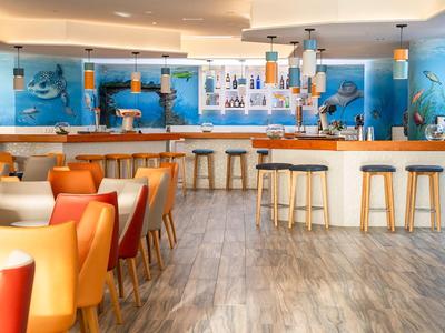 Bar d'hôtel lumineux avec des chaises orange et blanches et des tabourets en bois au comptoir.