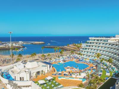 Gran hotel con piscina, tumbonas y vista al mar azul bajo cielo despejado.
