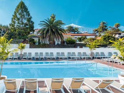 Grande piscina all'aperto con lettini e palme davanti a un edificio dell'hotel sotto un cielo azzurro.