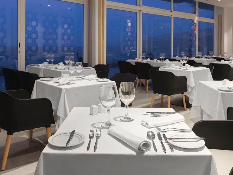 Elegant restaurant met witte tafelkleden, gedekte tafels en zwarte stoelen naast grote ramen.