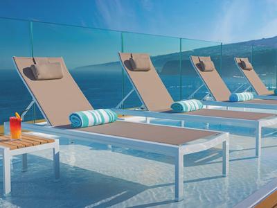 Quattro lettini con asciugamani e drink vicino all'acqua con vista sulle montagne.