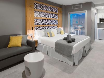 Habitación de hotel moderna con cama doble, sofá gris, mesa blanca y vista a balcón exterior.