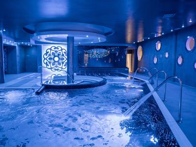 Modernes Hallenbad mit blau beleuchtetem Whirlpool und dekorativen Wandfenstern