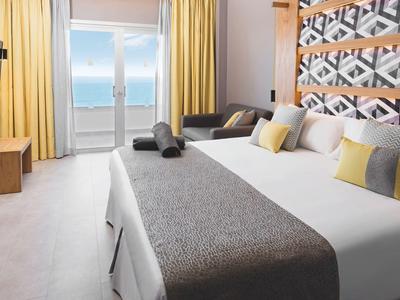 Helles Hotelzimmer mit großem Bett, dekorativen Kissen, Sofa und Meerblick durch große Fenster.