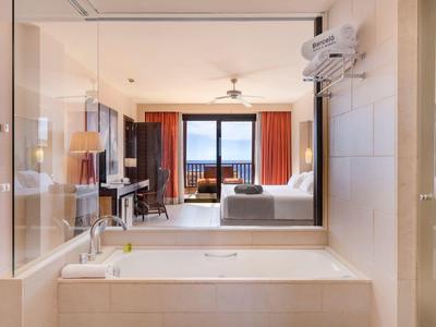 Moderne Badezimmereinrichtung mit großer Badewanne, Fensterblick auf Wohnzimmer und Meer dahinter.