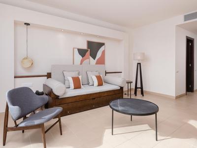 Habitación de hotel amueblada modernamente con cama, sillones y mesa redonda sobre suelo claro