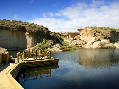 Wooden dock on calm lake surrounded by rocky cliffs under a partly cloudy blue sky.