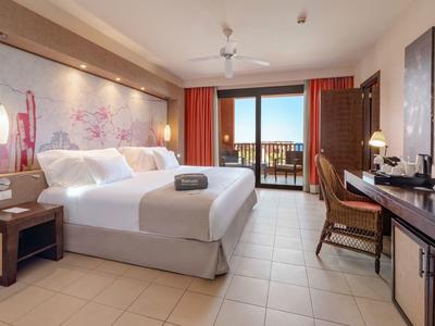 Chambre d'hôtel lumineuse avec grand lit, bureau, chaise, balcon et éclairage chaleureux.