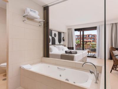 Bright hotel room with bathtub, double bed, and balcony overlooking neighboring buildings.