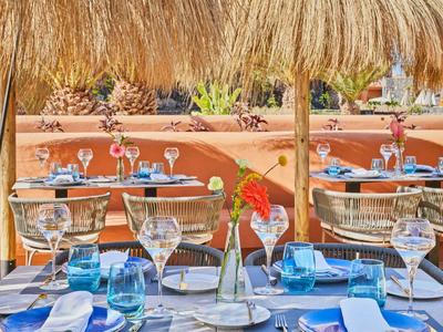 Outdoor restaurant seating with tables set under straw umbrellas and desert plants in the background.