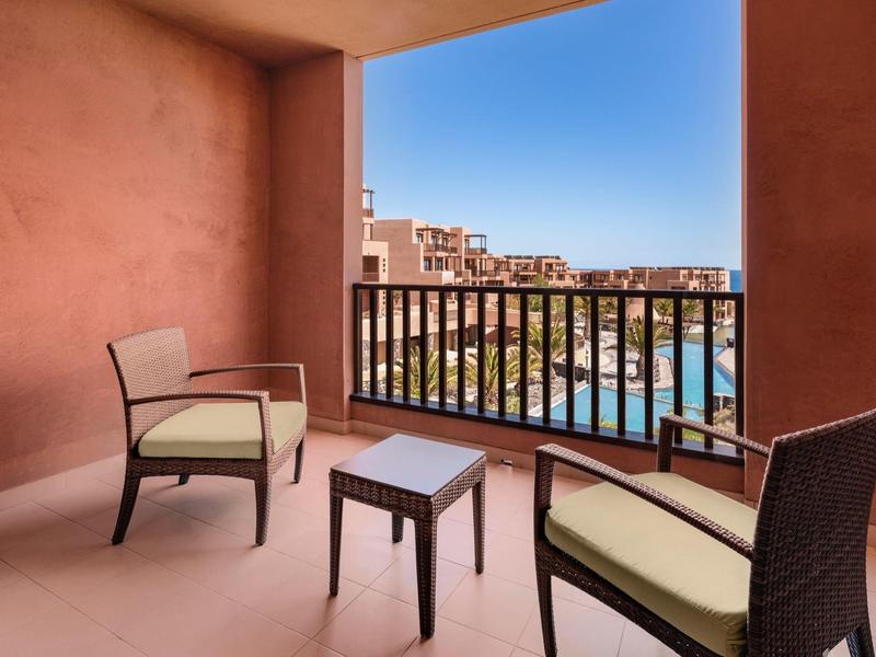 Balcony with two chairs and a table overlooking a resort pool and buildings under a clear blue sky.