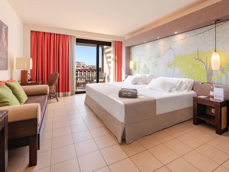 Modern hotel room with large bed, sofa, balcony view, and soft lighting.