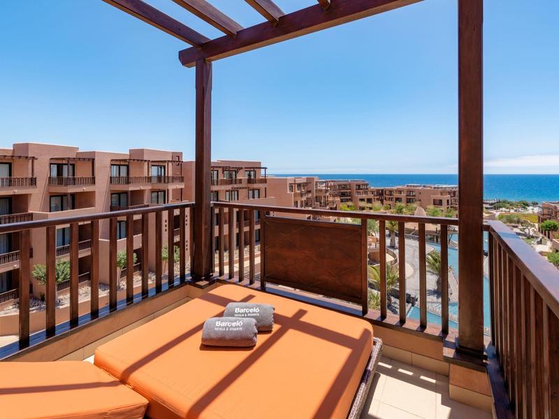 Balcony with orange lounge chairs overlooks buildings and sea under a clear blue sky.