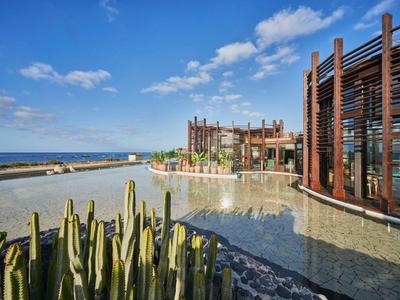 Complejo hotelero moderno junto al agua con cielo despejado y cactus en primer plano.