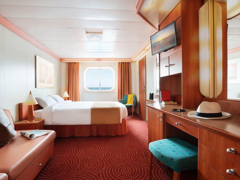 Bright cruise ship cabin with a bed, round window, and wooden furnishings.