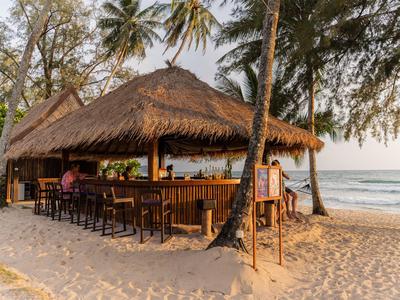 Offene Strandbar mit Strohdach und Barhockern am Sandstrand neben Palmen.