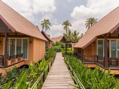 Holzsteg führt zu Bungalows zwischen üppigem Grün und Palmen unter bewölktem Himmel.