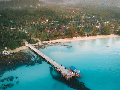 Langer Holzsteg führt über türkisfarbenes Wasser zu einem tropischen Strand mit Wald.