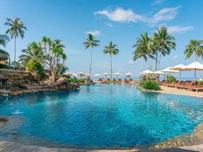 Large pool with sun loungers, umbrellas, and palm trees at a sunny vacation spot.
