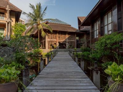 Ein Holzsteg führt durch einen tropischen Garten zu traditionellen Holzhäusern unter blauem Himmel.