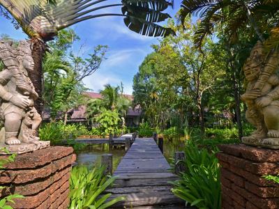Tropischer Garten mit Steg, umgeben von üppiger Vegetation und zwei steinernen Statuen.