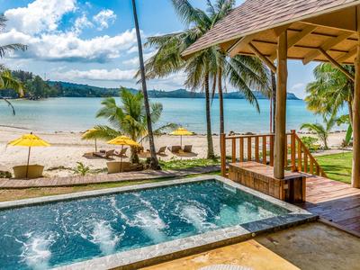 Blick auf einen Pool und einen ruhigen Sandstrand mit Palmen und klarem Himmel.