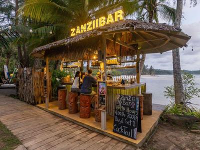 Offener Cocktailbar am Wasser mit Palmen und Holzstühlen, beleuchtet bei Dämmerung.