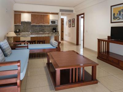 Zona giorno con mobili in legno, divano, tavolino e cucina sullo sfondo in una camera d'hotel