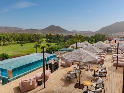 Moderna terraza de hotel con piscina, áreas de descanso y vistas a paisaje verde y montañas.