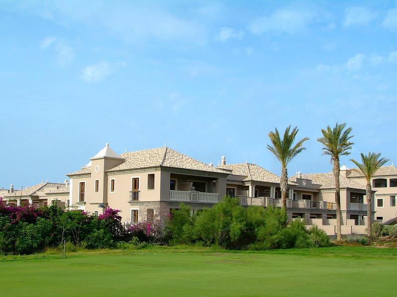 Complesso alberghiero moderno con palme davanti a un prato verde ben curato sotto un cielo azzurro.