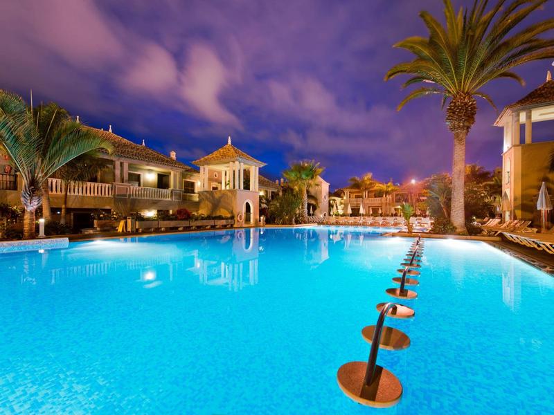 Area piscina illuminata di un hotel di notte con palme e posti a sedere in acqua.