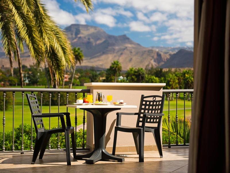 Balcone con tavolo, sedie e bevande con vista su palme e montagne.