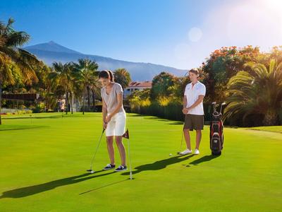 Zwei Personen beim Golfspielen auf einem grünen Platz mit Palmen und Bergen im Hintergrund bei Sonnenschein.