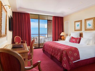 Helles Schlafzimmer mit großem Bett, roten Vorhängen, Teppich und Balkon mit Meerblick.
