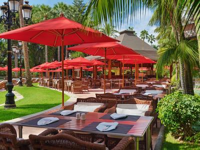 Outdoor-Bereich eines Restaurants mit roten Sonnenschirmen, Holztischen und grünem Palmenbewuchs.