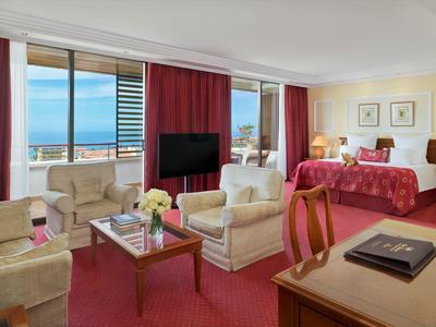 Helles Hotelzimmer mit Sitzbereich, rotem Bett, Balkon und Meerblick bei Tageslicht.