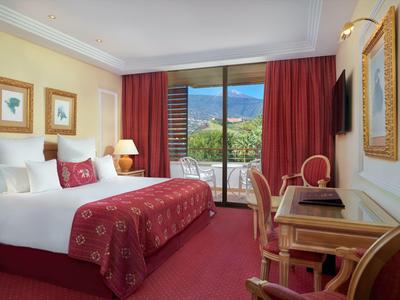 Gemütliches Hotelzimmer mit Doppelbett, rotem Teppich, Schreibtisch und Blick auf Balkon und Berge.