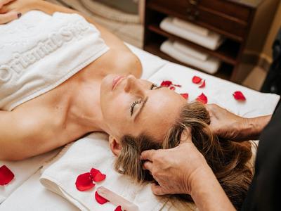 Frau entspannt bei Kopfmassage auf weißem Handtuch mit roten Rosenblättern.