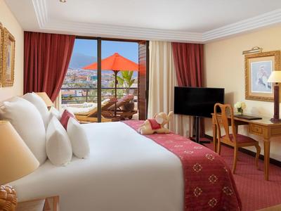 Gemütliches Hotelzimmer mit großem Bett, rotem Teppich, Schreibtisch, TV und Balkon mit rotem Sonnenschirm.
