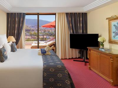Helles Hotelzimmer mit großem Bett, TV auf Kommode, offenem Balkon und Bergen im Hintergrund.