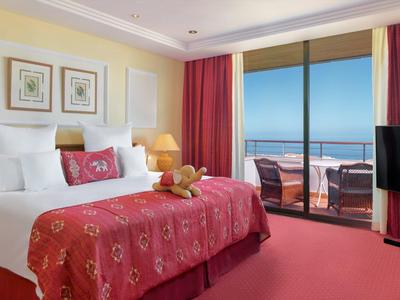Hell eingerichtetes Hotelzimmer mit Doppelbett, rotem Teppich, Balkon und Meerblick.